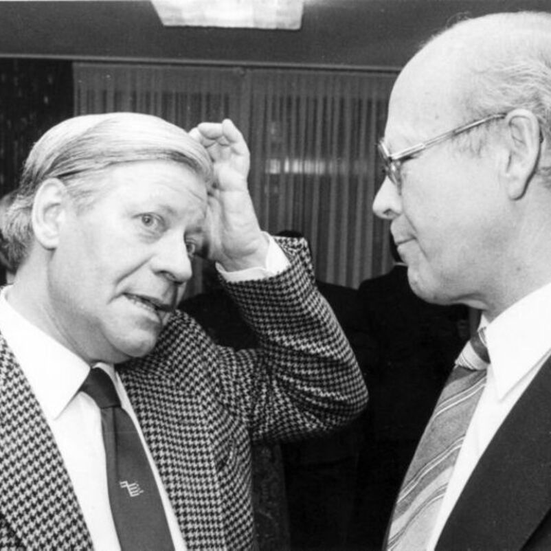 Das Bild zeigt zwei männlich lesbare Personen einander gegenüber stehend im Gespräch.