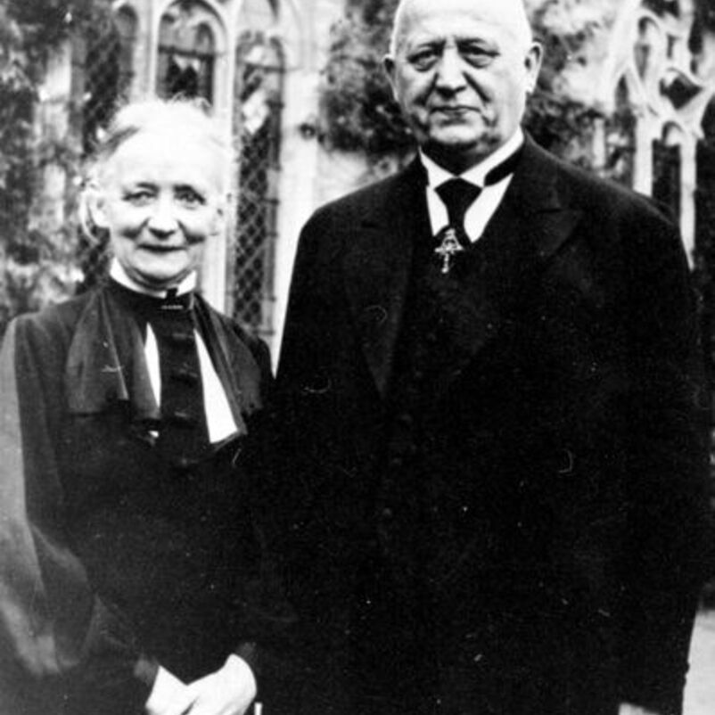 Das Schwarzweiß-Bild zeigt je eine männlich und eine weiblich lesbare Person, er in einem schwarzen Anzug, sie in einem dunklen Kleid, außen vor einer Kirchenmauer.