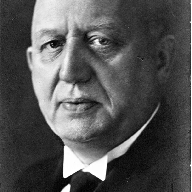 Das Schwarzweiß-Bild ist die Portraitaufnahme einer älteren männlich lesbaren Person mit Glatze, weißem Hemd und schwarzem Anzug.