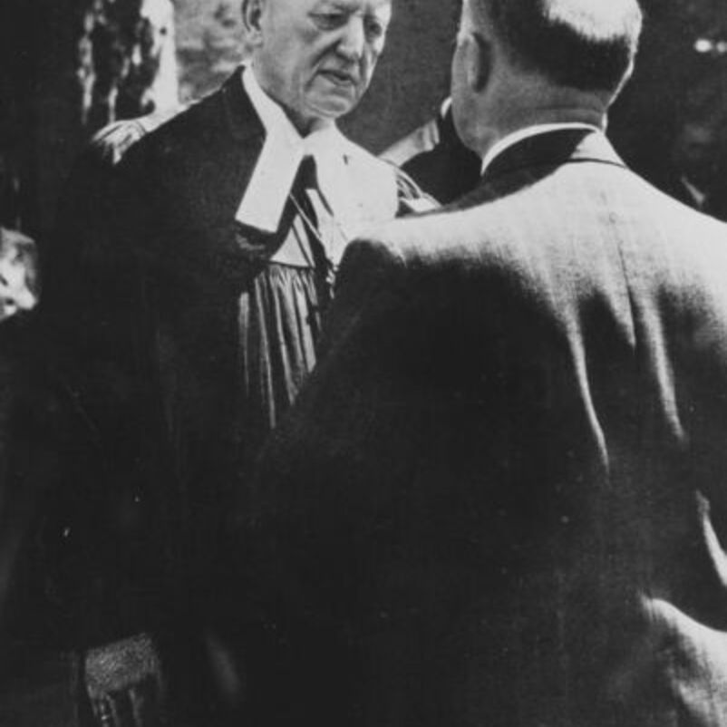 Das Schwarzweiß-Bild zeigt zwei männlich lesbare Personen, einer davon deutlich als Geistlicher erkennbar anhand seines Talars, die einander gegenüber stehen.