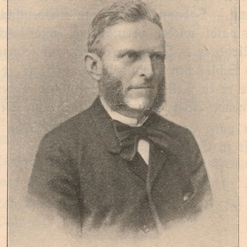 Das Bild zeigt eine schwarzweiße Aufnahme mit einer männlich lesbaren Person mit kurzen welligen Haaren, einem dichten Backen- und Kinnbart, einem weißen Hemd, einer dunklen gebundenen Schleife um den Hals und ein einem dunklen Sakko.