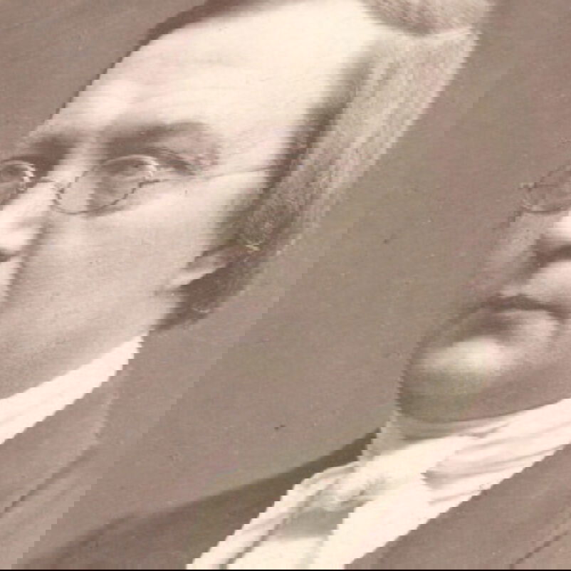 Der Bild zeigt eine schwarzweiße Aufnahme einer männlich lesbaren Person ohne Bart, mit kurzen mittelbloden Haaren, einer kleinen Brille mit Metallfassung, einem weißen Hemd, einer gebundenen Schleife um den Hals und einem dunklen Sakko.