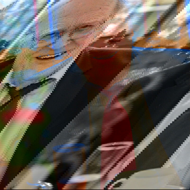 Eine männlich lesbare Person mit weißen Haaren und Brille, weißem Hemd, dunklem Sakko und dunkler Krawatte.