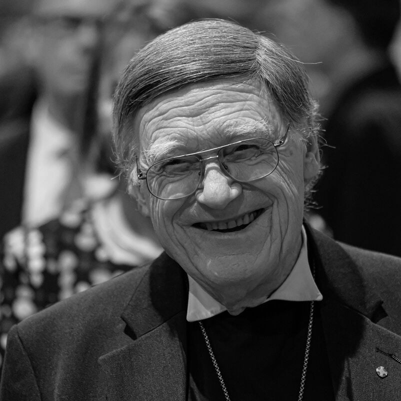 Eine männlich lesbare Person mit dunklen Haaren und Brille, lachend, in schwarzweiß.