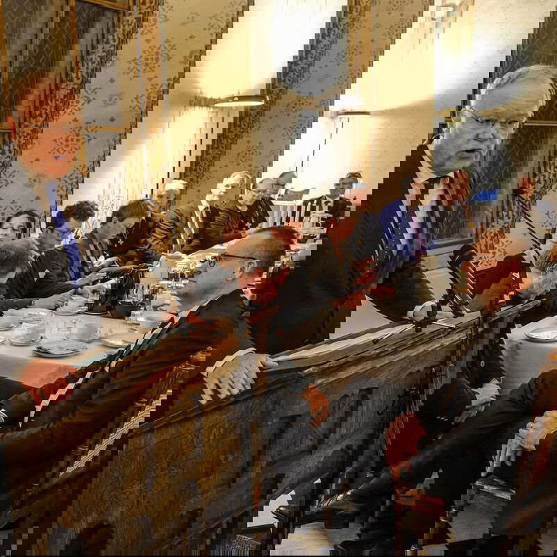 Ein Mann im Anzug mit blauer Krawatte steht an einem Rednerpult. Links von ihm sitzen etwa ein Dutzend Menschen an einem gedeckten Tisch.