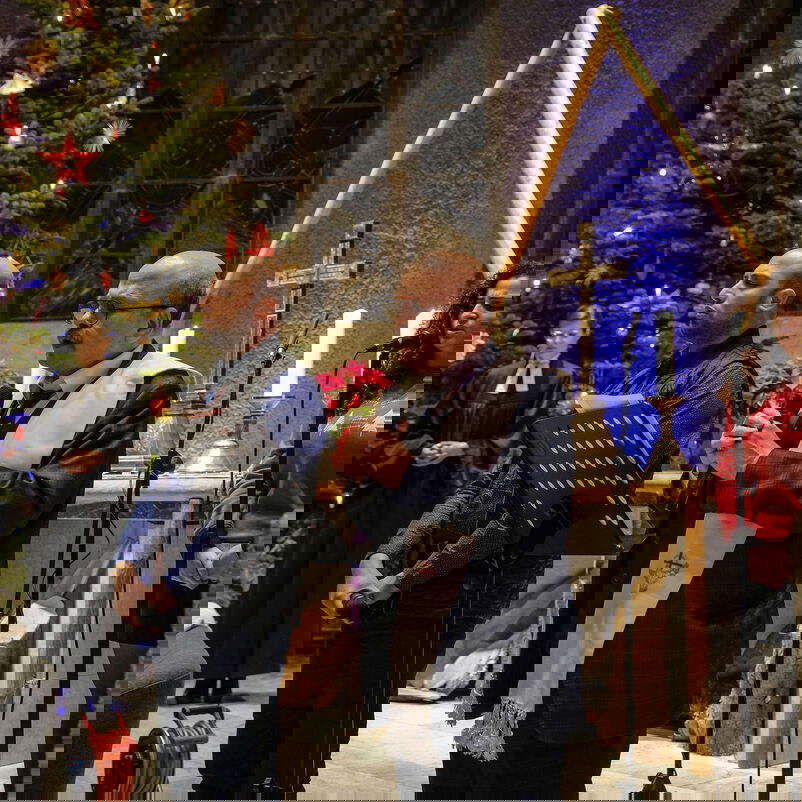 Vier Personen stehen an Mikrophonen, einer liest von einem Notenständer. Im Hintergrund stehen ein großer Weihnachtsbaum und ein Kreuz.