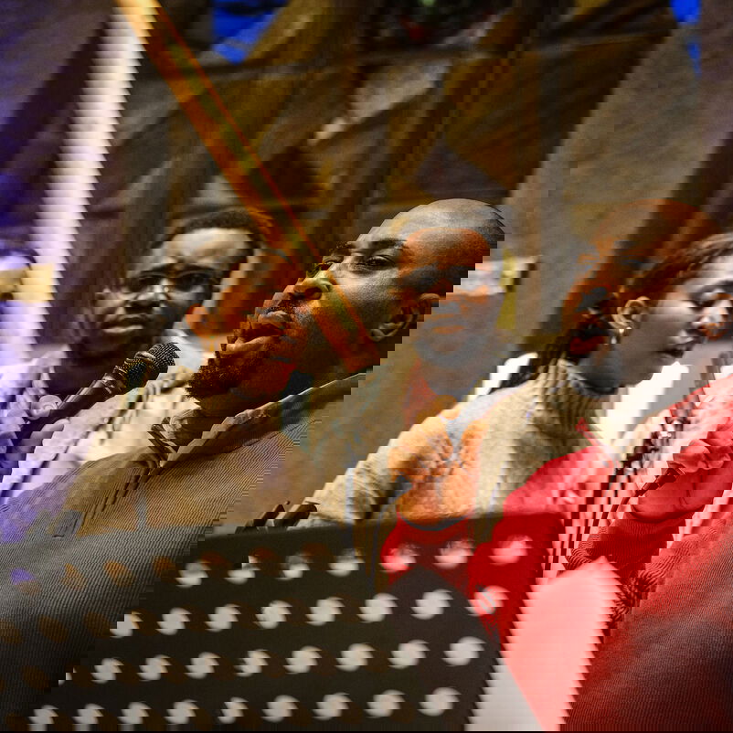 Drei Personen, eine mit leuchtend rotem Pulli und Mikro in der Hand, stehen zusammen vor einem Kreuz in einer Kirche.