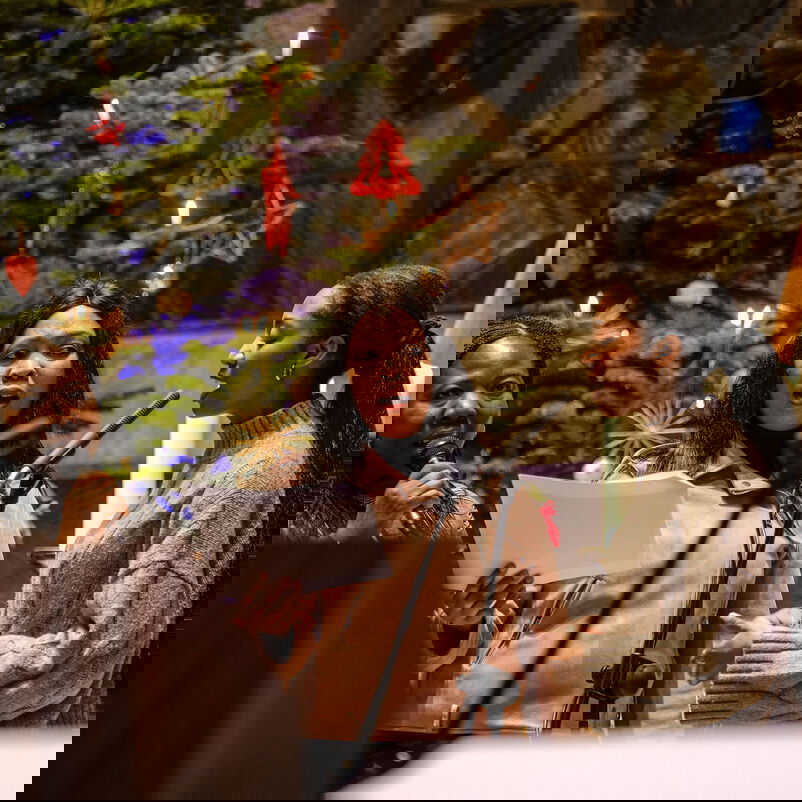 Drei Frauen mit schwarzen Haaren singen in Mikros, vor einem geschmückten Weihnachtsbaum.