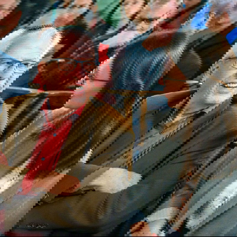 Eine ältere, männlich gelesene Person mit Kippa auf dem Kopf und eine jüngere, weiblich gelesene Person, sind sich im Gespräch zugewandt, auf Stühlen vor weiteren Menschen sitzend.