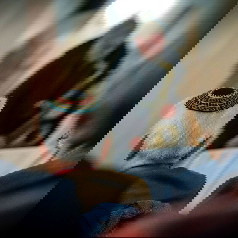 Eine Person mit kurzen, weißen Haaren und einer Kippa ist zwischen anderen Personen hindurch von hinten, in einer Kirchenbank sitzend, zu sehen.