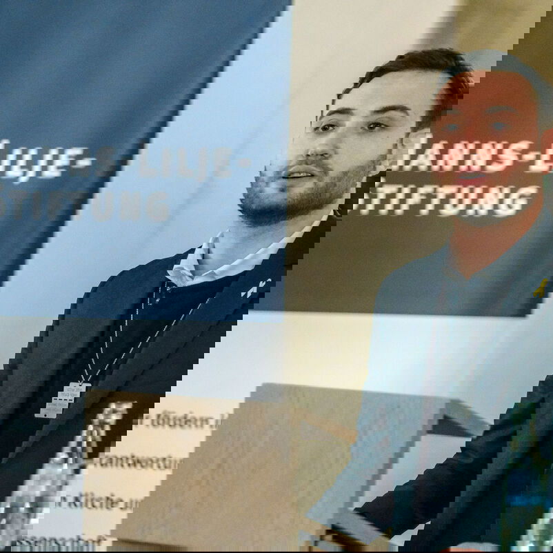 Eine männlich gelesene Person mit dunklem Pulli und Jackett an einem Redepult. Im Hintergrund ein blau-weißer Aufsteller mit dem Schriftzug "Hanns-Lilje-Stiftung".