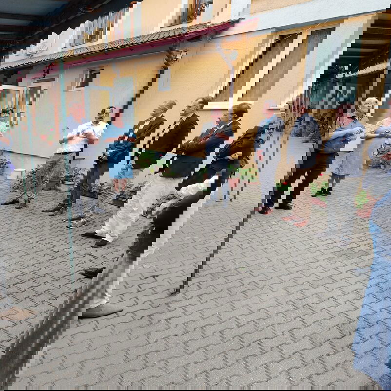 Eine Gruppe von weiblich und männlich zu lesenden Personen steht im Kreis vor einem Haus und unterhält sich.