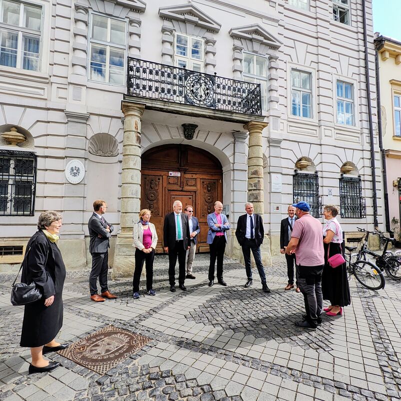 Eine Gruppe von weiblich und männlich zu lesenden Personen steht auf einem Platz vor einem Gebäude.