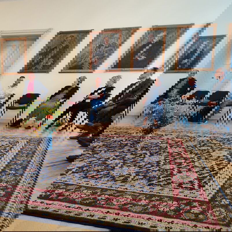 Eine Gruppe von weiblich und männlich zu lesenden Personen sitzt in einem Halbkreis um einen Teppich herum und spricht miteinander.