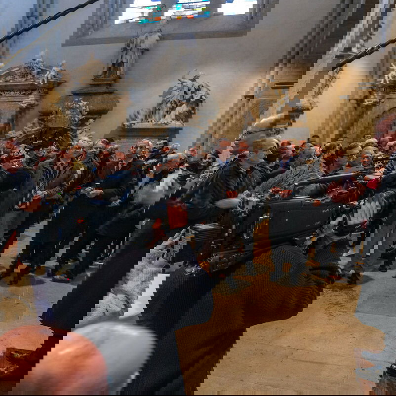 Eine männlich zu lesende Person steht in einer Kirche vor einer Säule und spricht zu einer Gruppe von Menschen.