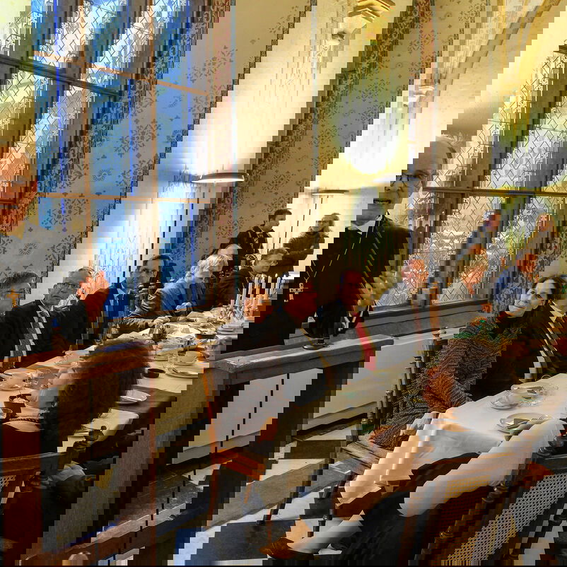 Eine männlich zu lesende Person steht an einem Redepult und spricht zu Menschen, die an einem Tisch sitzen.