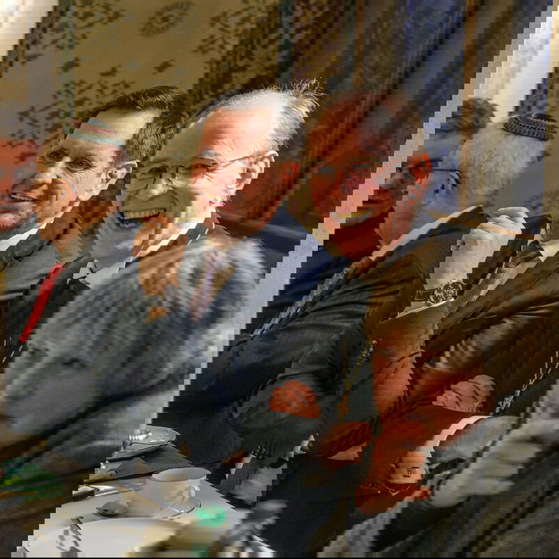 Vier männlich und eine weiblich gelesene Person sitzen nebeneinander an einem gedeckten Tisch, zwei lachen herzlich. 