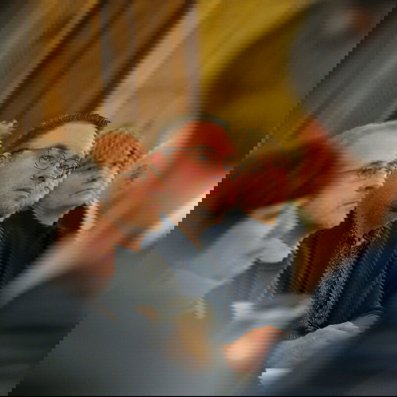 Männlich und weiblich zu lesende Person sitzen und hören einer Rede zu.