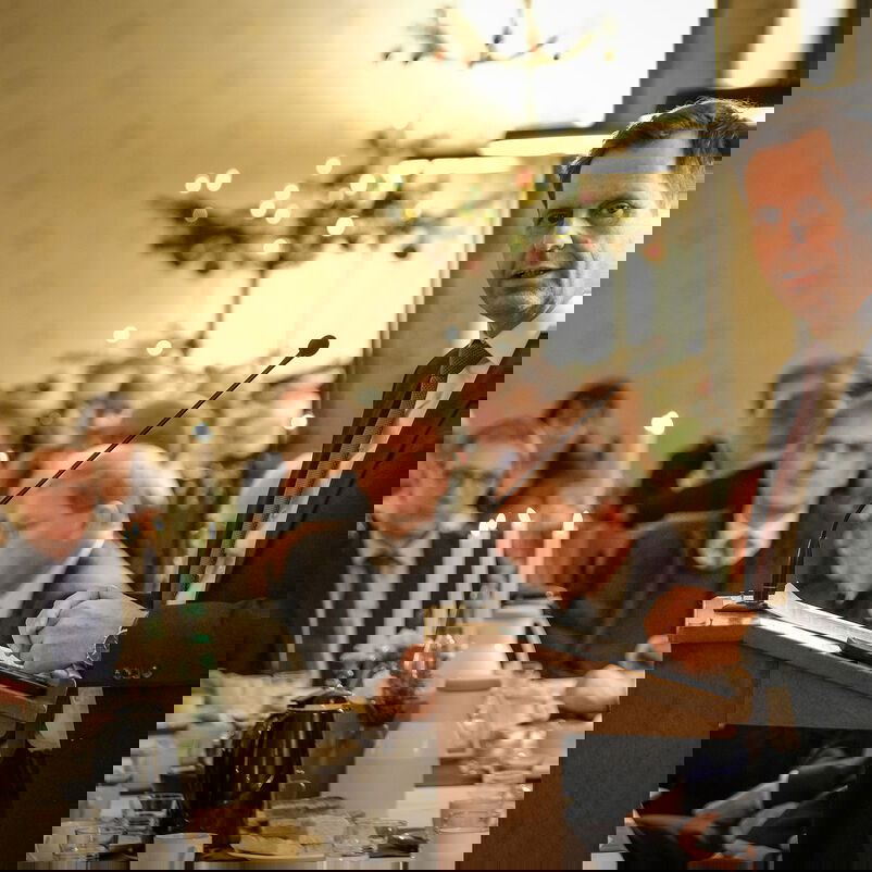 Eine männlich zu lesende Person steht an einem Redepult und spricht zu Menschen, die an einem festlich gedeckten Tisch sitzen.