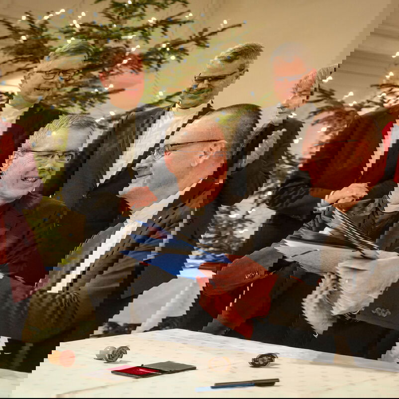 Zwei männlich zu lesende Personen sitzen an einem Tisch und tauschen lächelnd einen unterschrieben Vertrag aus. Im Hintergrund sind weiblich und männlich zu lesende Personen zu sehen.