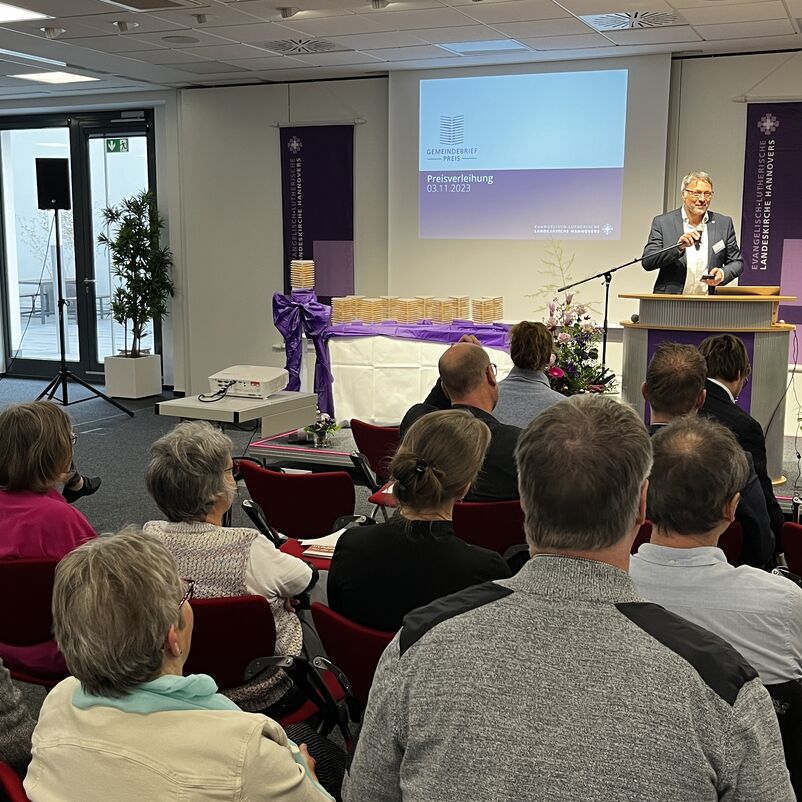 Eine männlich lesbare Person am Rednerpult auf einer Bühne, davor viele Menschen von hinten auf Stühlen sitzend.