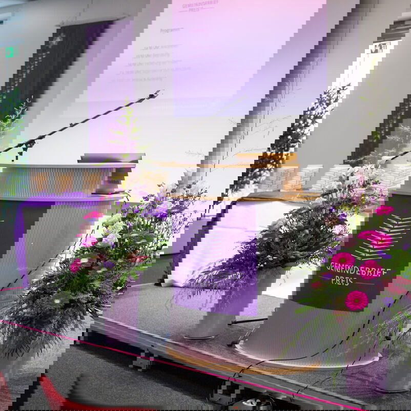 Auf einer Bühne steht ein Redepult, das gerahmt wird von Blumensträußen.