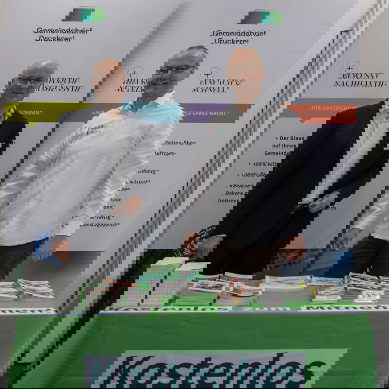 Zwei männlich zu lesende Personen stehen hinter einem Tisch mit Broschüren. Vom Tisch hängt ein Transparent mit der Aufschrift "Kostenlos Mitnehmen!"