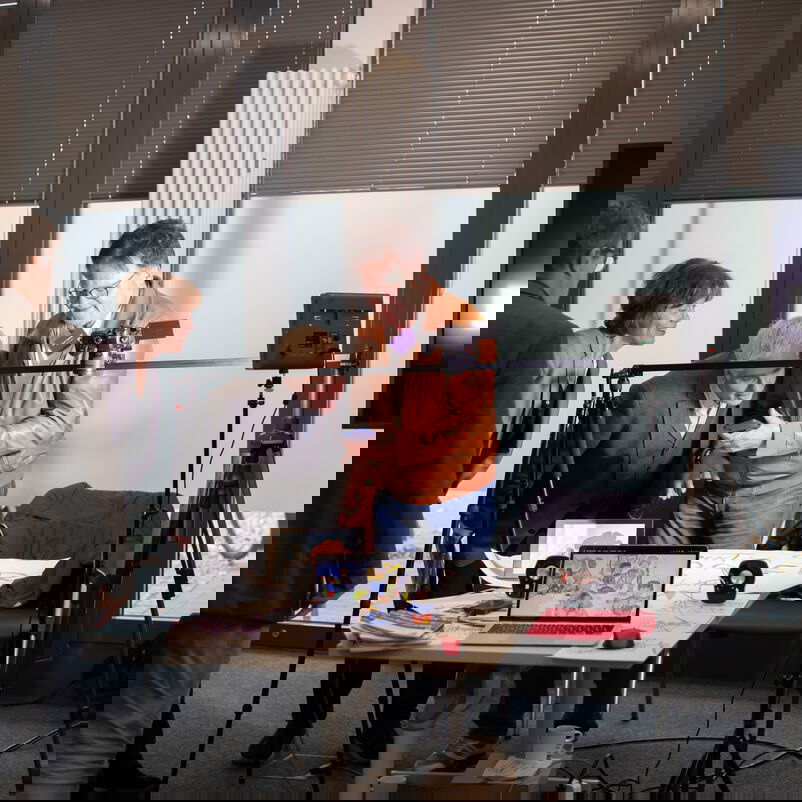 Eine Gruppe von Menschen steht um einen Tisch herum. Eine männlich zu lesende Person zeigt etwas auf dem Handy, das von den anderen betrachtet wird.