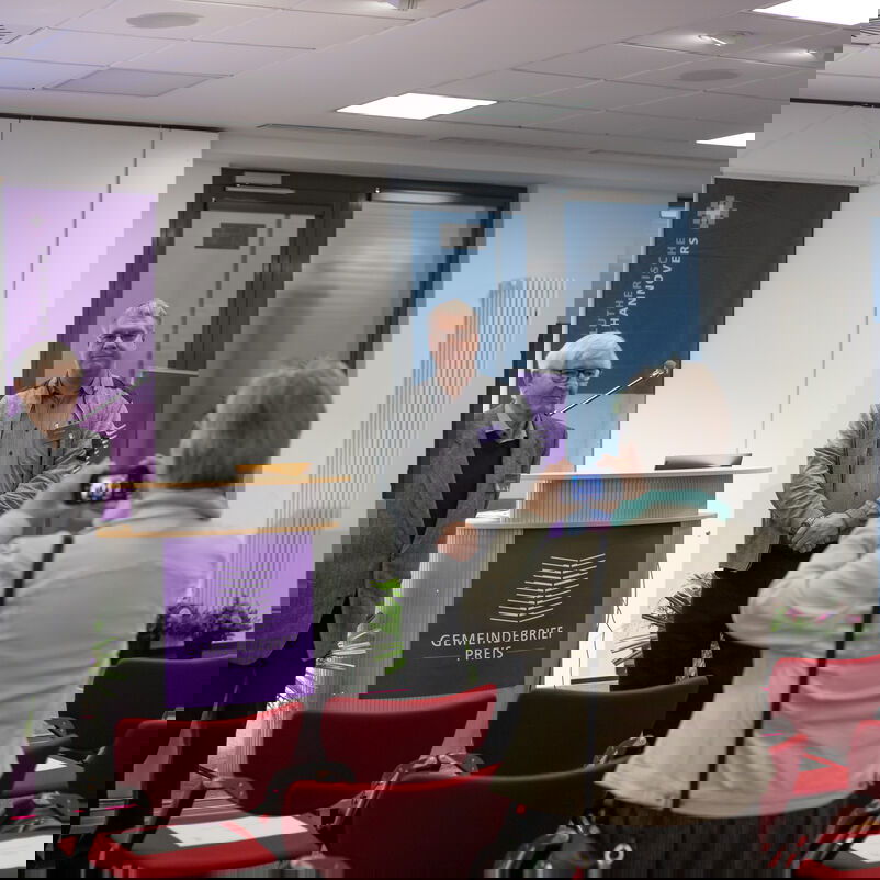 Drei Menschen stehen verteilt um das Rednerpult der Gemeindebriefpreisverleihung. Eine vierte Person macht ein Foto von ihnen.