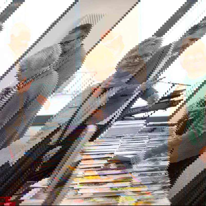 Männlich und weiblich zu lesende Personen schauen sich ausliegende Informationsmaterialen an und unterhalten sich.