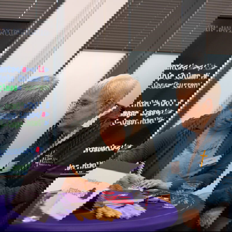 Zwei weiblich zu lesende Personen stehen an einem Stehtisch, auf dem Materialien ausliegen. Eine der Personen schreibt etwas auf, die andere schaut zu.