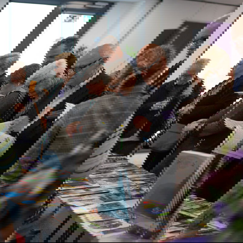 Männlich und weiblich zu lesende Personen schauen sich Infomaterial an, das auf Tischen ausliegt.