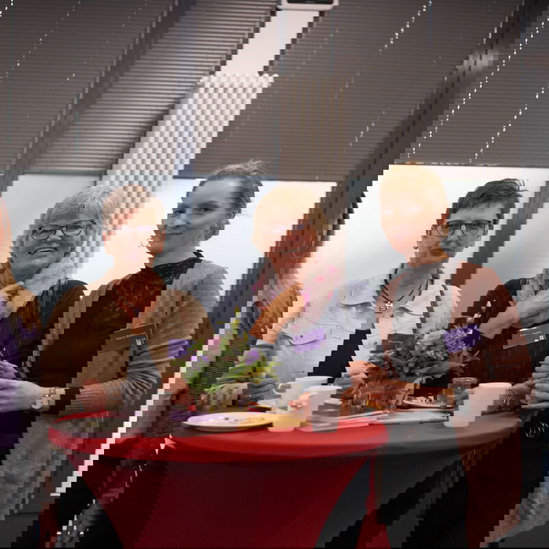 Um einen Tisch stehen vier weiblich zu lesende Personen und lächeln den Betrachter an.