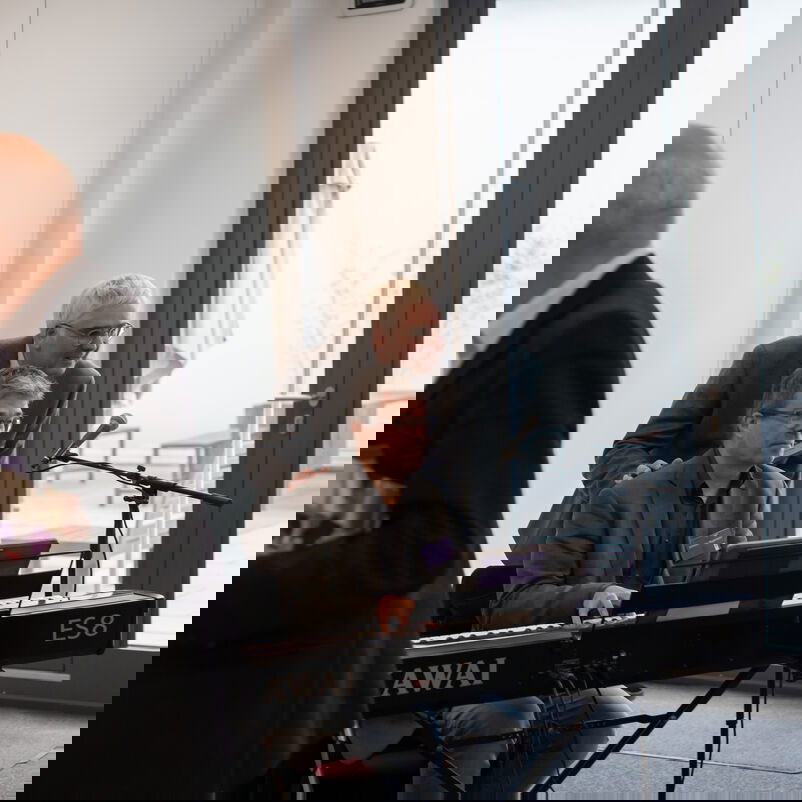 An einem E-Piano sitzt eine männlich zu lesende Person. Eine weitere Person männlich zu lesende Person steht hinter dem Pianisten und beugt sich vor. Beide blicken zu einem Fotografen, der sie fotografiert.