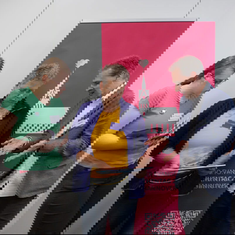 Drei Personen stehen vor einem Plakat, das eine weiße, stilisierte Kirche mit Herz über dem Dach vor rosafarbigem Hintergrund zeigt. Einer Person berät die anderen zwei. Alle betrachten einen Flyer in den Händen der mittigen Person.