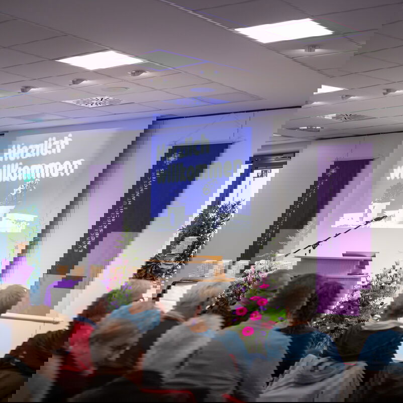 Durch einen Beamer wird die Begrüßung "Herzlich Willkommen" an die Wand hinter einem Rednerpult geworden. Davor sind voll besetzte Stuhlreihen zu sehen.