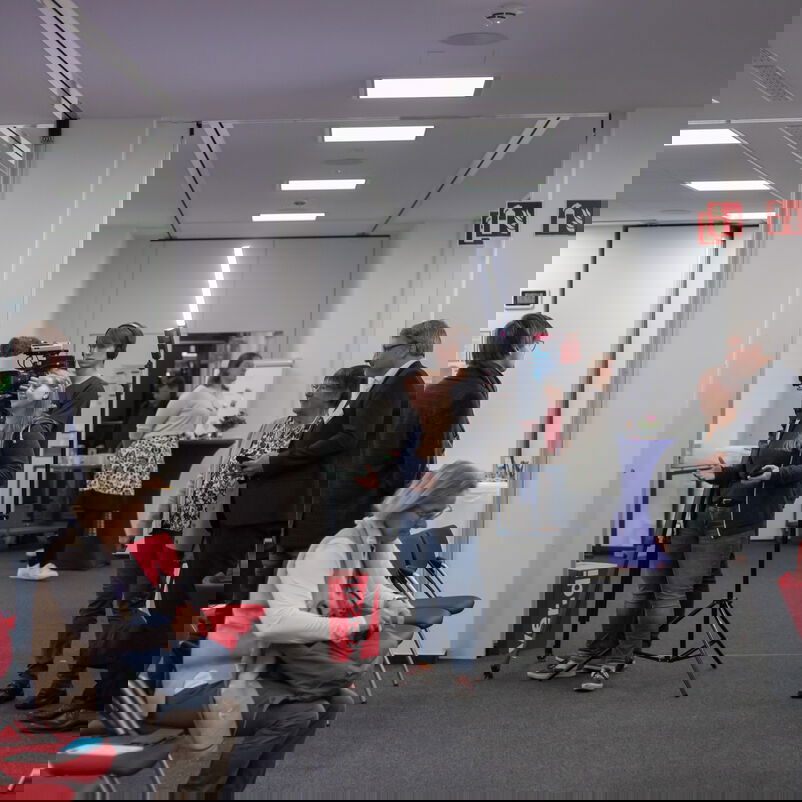 In einem Raum stehen zwei junge Leute mit Videokamera und Lichttechnik im Interview mit einer weiteren Person. Einige der umgebenden Personen verfolgen das Interview.