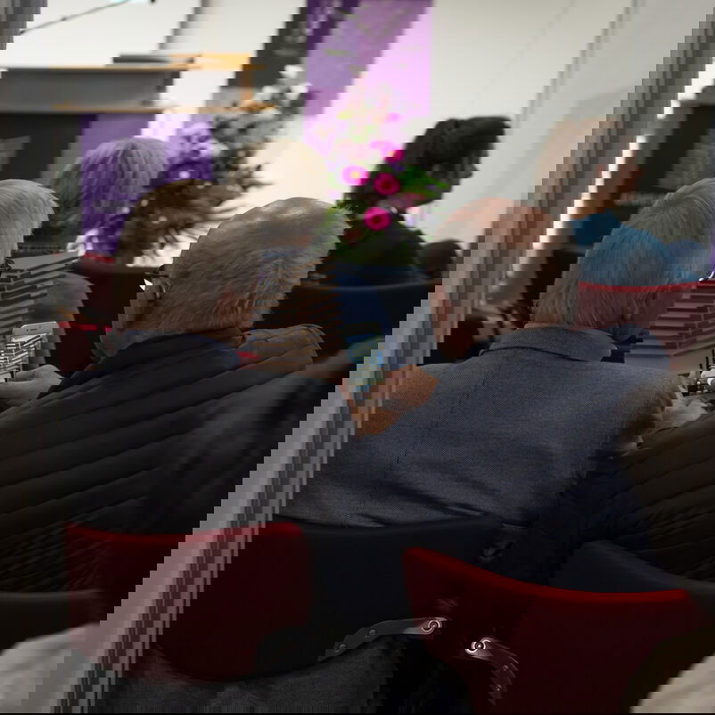 Zwei ältere männlich zu lesende Personen sitzen im Publikum mit dem Rücken zur betrachtenden Person. Mit dem Handy fotografieren sie ihren Pokal.