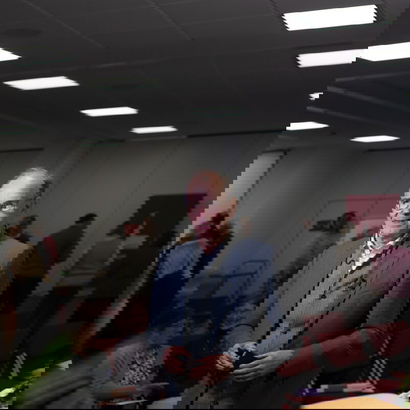 Eine männlich zu lesende Person steht in einem Raum während eines Interviews. Im Hintergrund befinden sich weitere Personen im Raum verteilt.