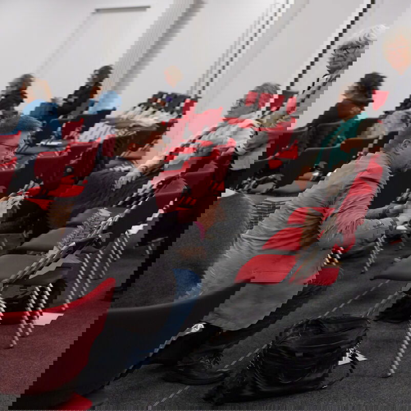 Eine männlich zu lesende Person hockt vor einem Stuhl in einer Stuhlreihe und fotografiert den darauf abgestellten Pokal.