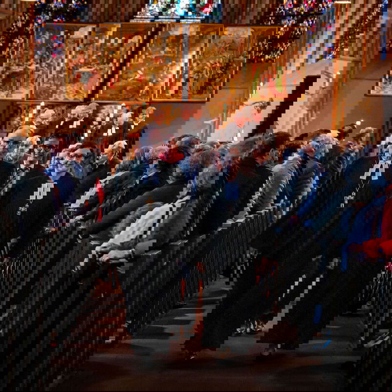 Eine männliche und eine weiblich zu lesende Person laufen in Talaren einen Mittelgang in einer Kirche entlang, sie führen eine Delegation an. Links und rechts stehen viele Menschen. Im Hintergrund leuchtet ein goldenes Altarbild.