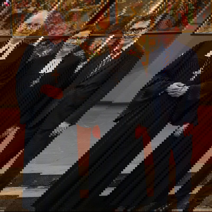 Zwei männlich zu lesende Personen und eine weiblich zu lesende Person stehen auf Treppenstufen. Im HIntergrund ist ein Altar zu sehen.