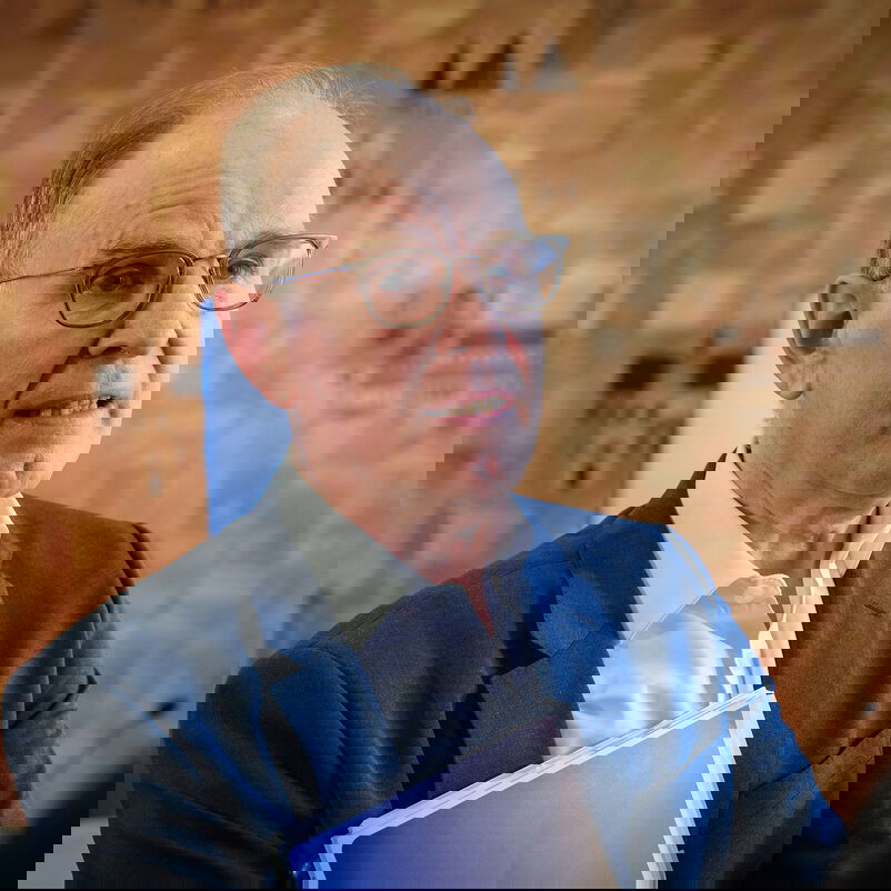 Eine männlich lesbare Person mit Brille und kurzen grauen Haaren, ohne Bart, mit blauem Sakko und weißem Hemd im Gespräch.