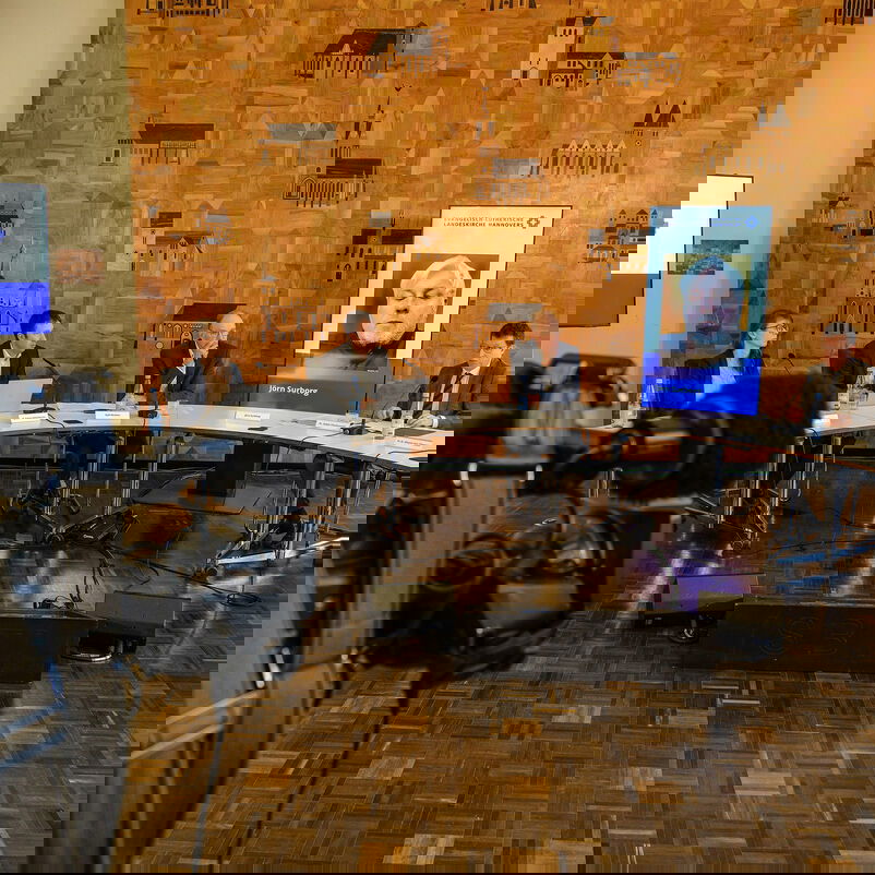 Ein Konferenzsaal mit einem Podium, hinter den Tischen sitzen Personen, im Vordergrund steht eine Fernsehkamera.