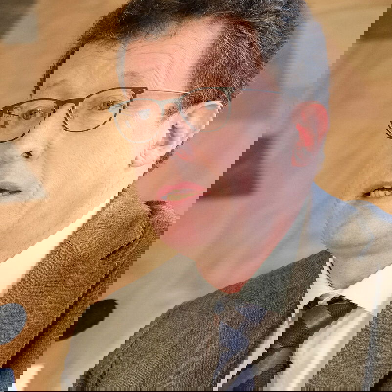 Eine männlich lesbare Person mit kurzen braunen Haaren, Brille, braunem Sakko und weißem Hemd im Gespräch.