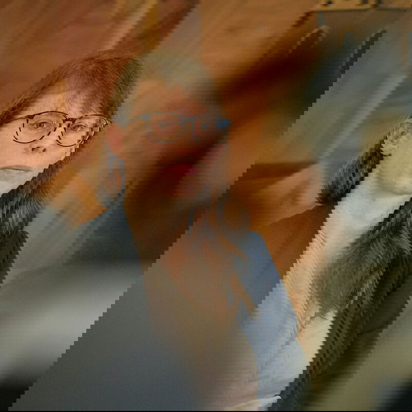 Eine weiblich lesbare Person mit langen Haaren und Brille sitzend am Podiumstisch.