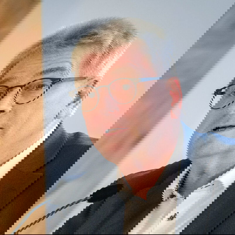 Eine männlich lesbare Person mit kurzen grauen Haaren, Brille und schwarzem Sakko und weißem Hemd im Gespräch.