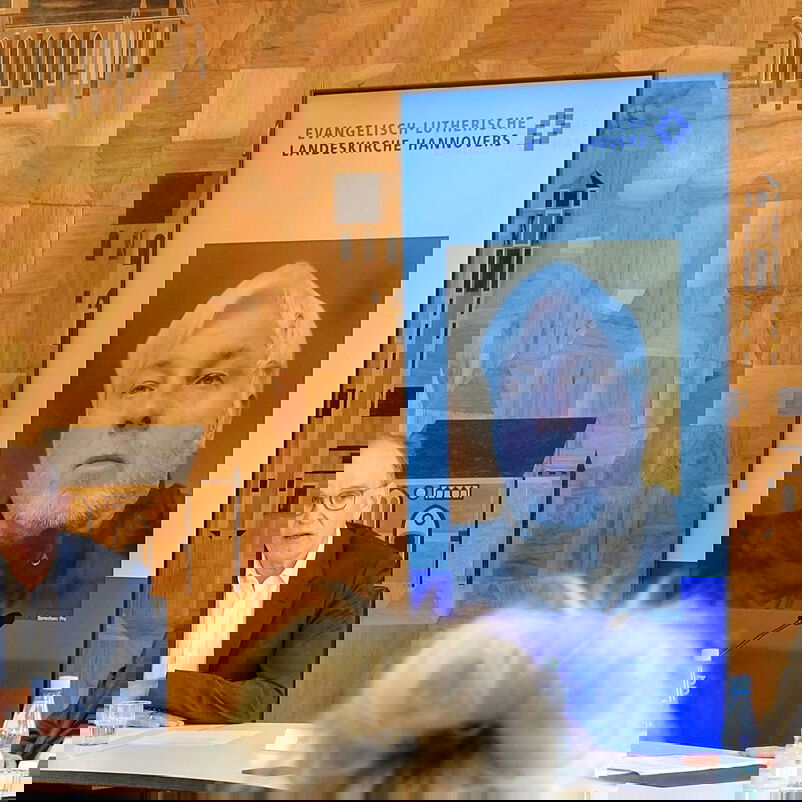 zwei männlich lesbare Personen sitzen an einem Diskussionspodium, eine dritte männlich lesbare Person ist über einen Bildschirm zugeschaltet.