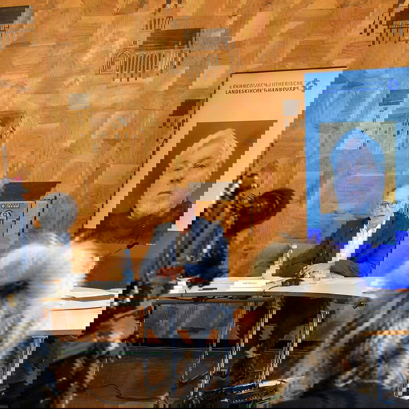 zwei männlich lesbare Personen am Podium, eine dritte männlich lesbare Person nimmt über einen Bildschirm am Podium teil, im Vordergrund steht eine Fernsehkamera.