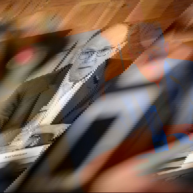 Eine männlich lesbare Person mit Brille und kurzen grauen Haaren, ohne Bart, mit blauem Sakko und weißem Hemd im Gespräch vor einer Kamera.